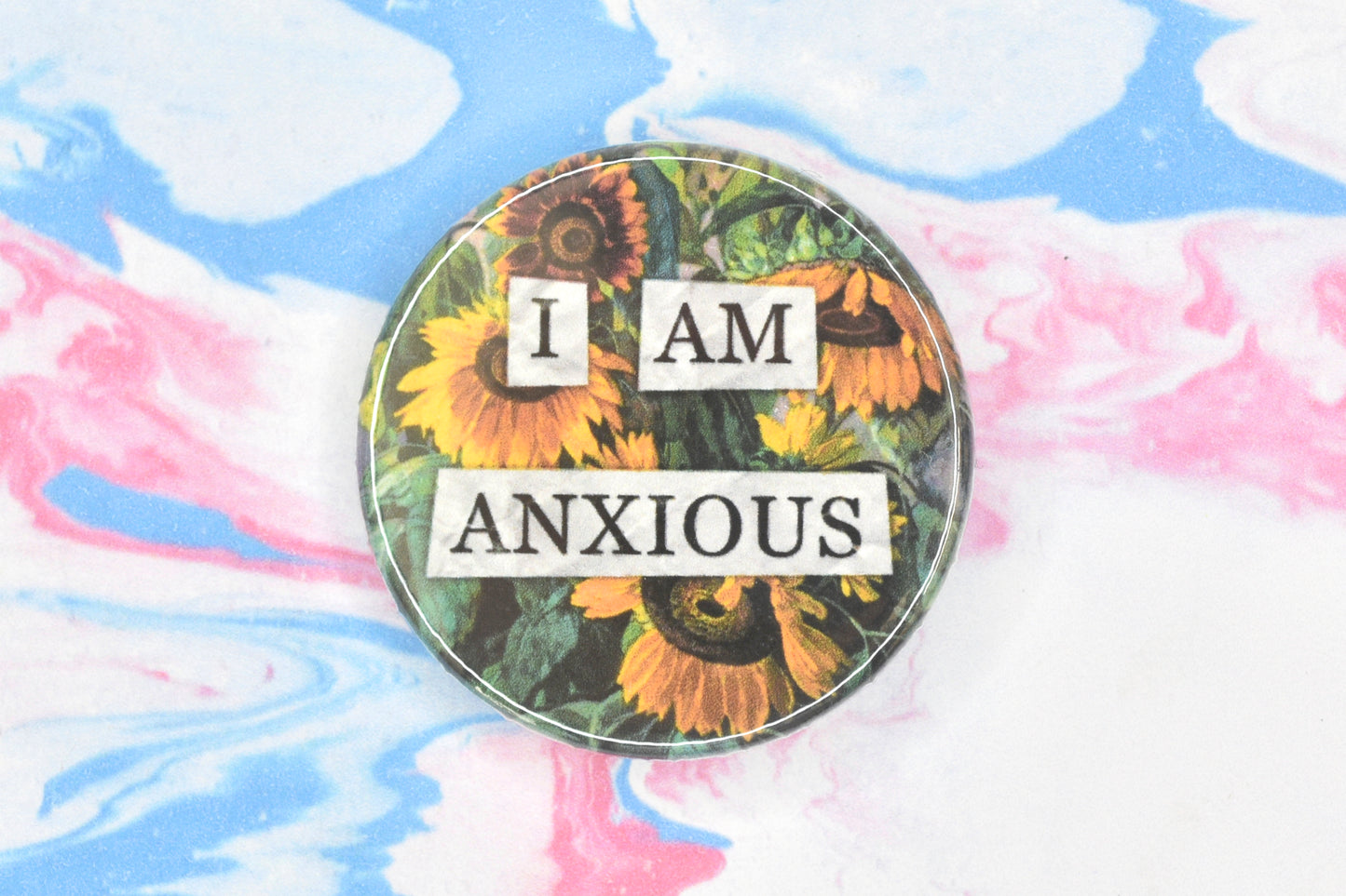 A circular button badge featuring a hidden disability sunflower design. Collaged text is overlaid on a painting of some sunflowers. The text reads "I am anxious".