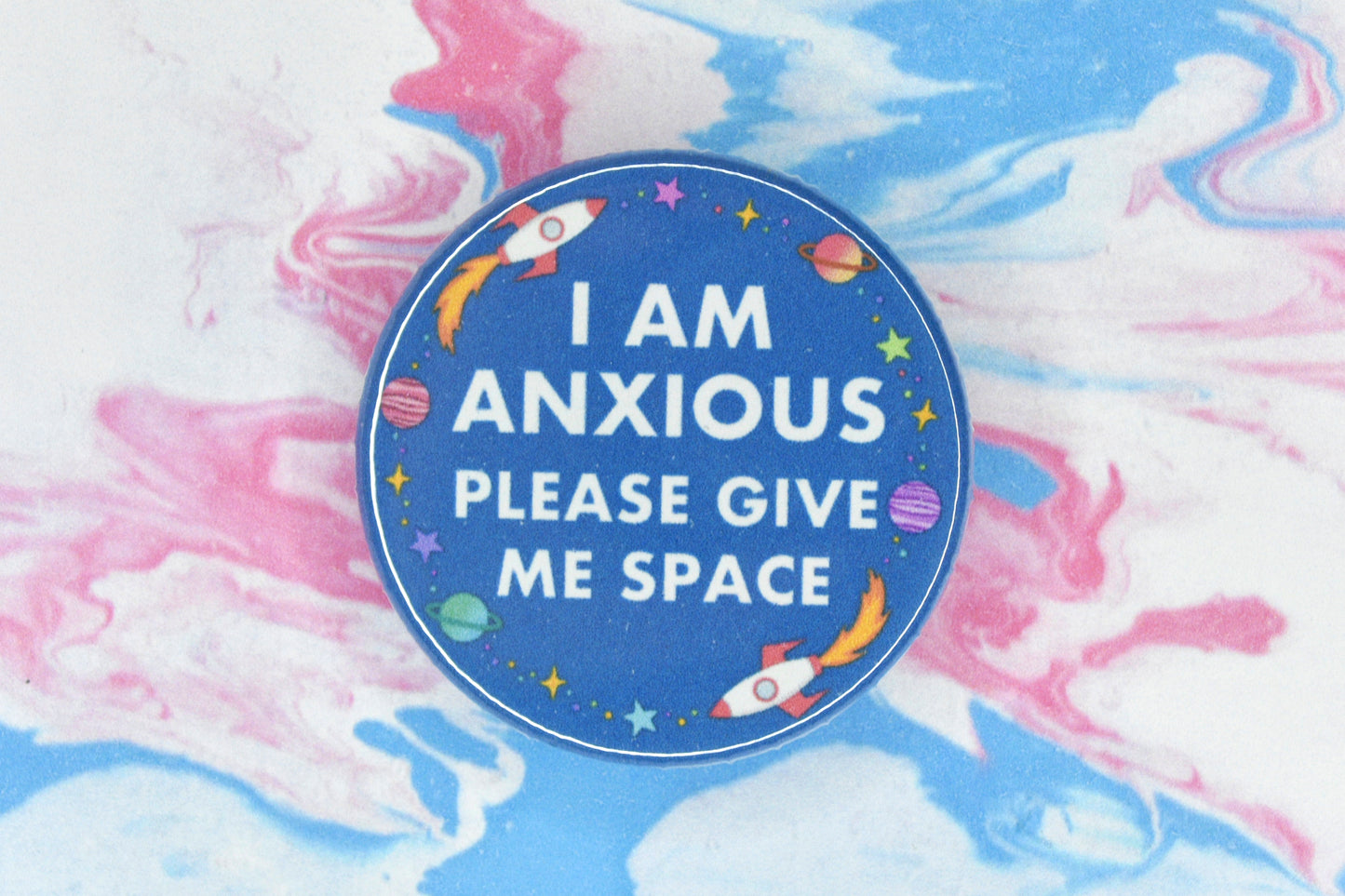 A hidden disability badge featuring a blue background and white text reading "I am anxious, please give me space". There are space related illustrations surrounding the text including rockets, planets and stars.