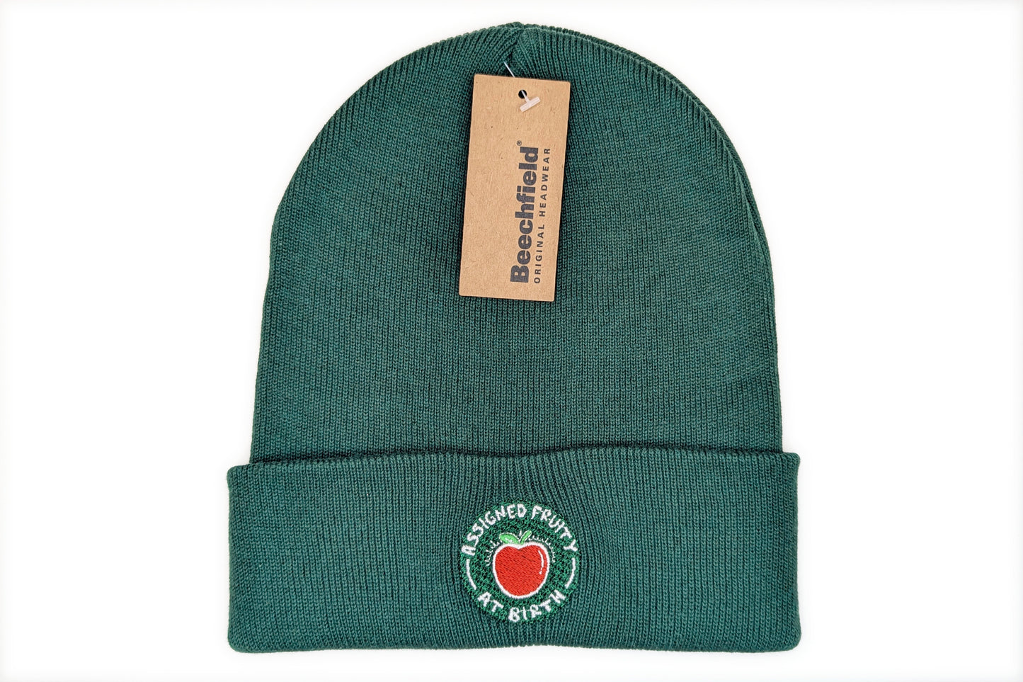 A photo of a bottle green cuffed beanie with an LGBTQ+ pride design embroidered on the cuff. The design depicts a red apple with white text surrounding it reading "Assigned Fruity At Birth"
