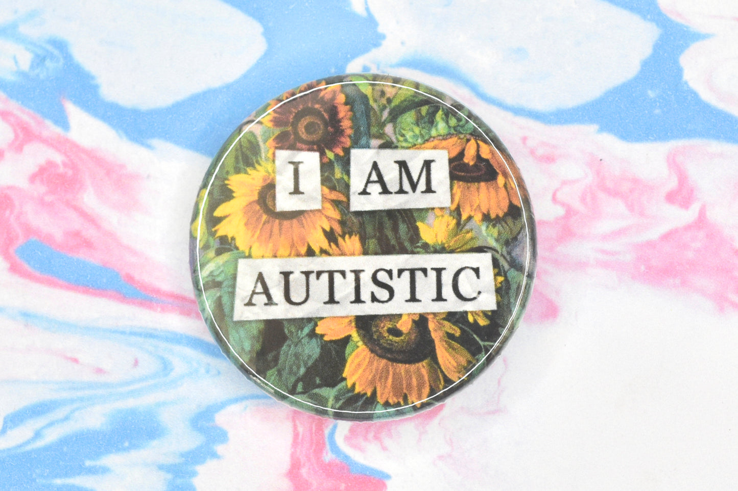 A circular button badge featuring a hidden disability sunflower design. Collaged text is overlaid on a painting of some sunflowers. The text reads "I am autistic".