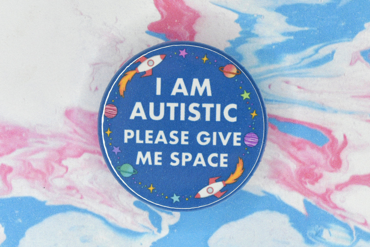 A hidden disability badge featuring a blue background and white text reading "I am autistic, please give me space". There are space related illustrations surrounding the text including rockets, planets and stars.