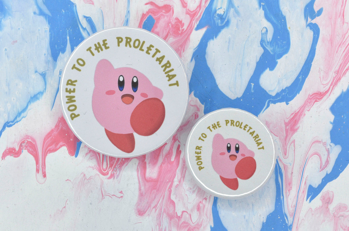 Two pin badges in sizes medium and small, both featuring an illustration of Kirby jumping on a white background. There is yellow/gold text in a curved line above the illustration which reads "power to the proletariat".