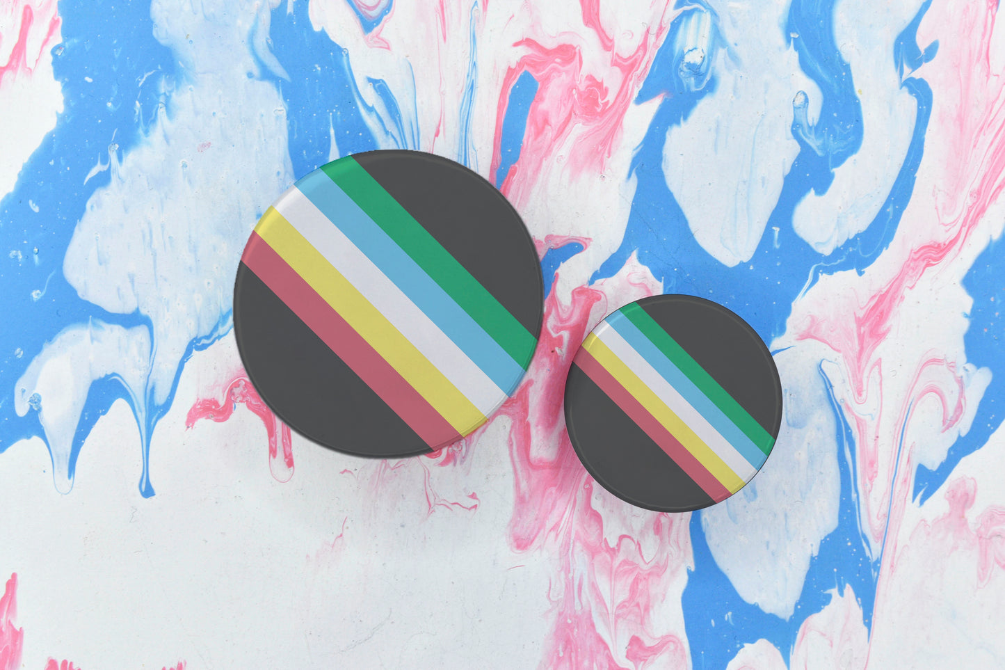 Two badges in sizes medium and small respectively, both featuring the disability pride flag, i.e. with red, yellow, white, blue and green bands running diagonally downwards on a dark grey background.