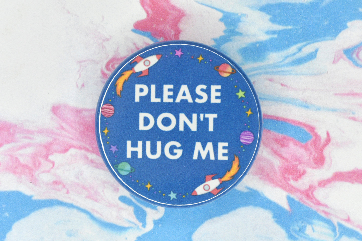 A hidden disability badge featuring a blue background and white text reading "Please don't hug me". There are space related illustrations surrounding the text including rockets, planets and stars.