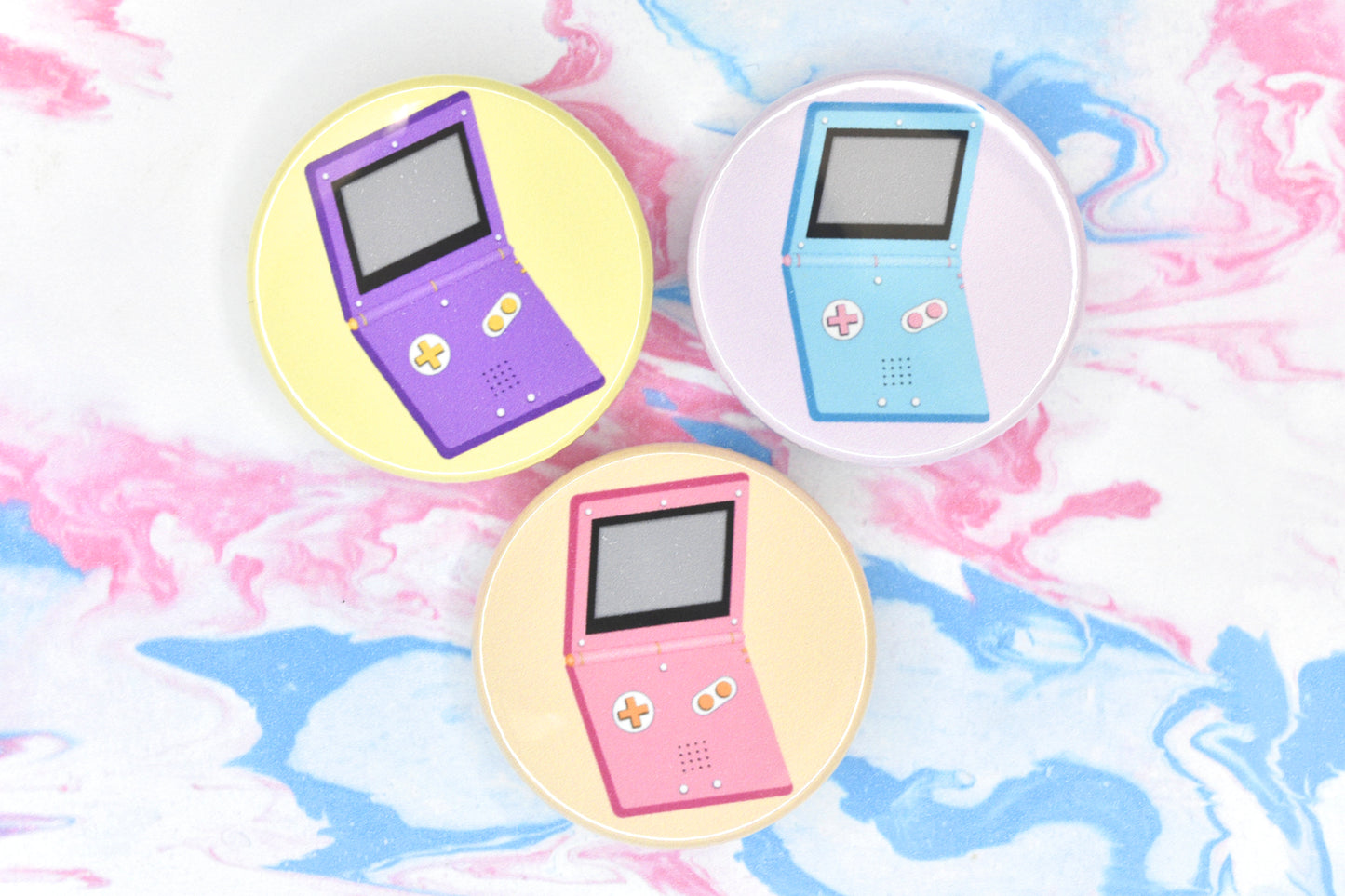 Three LGBTQ+ pride badges, each depicting a Gameboy Advance in different pride flag colours. The flags are non-binary, trans and lesbian.