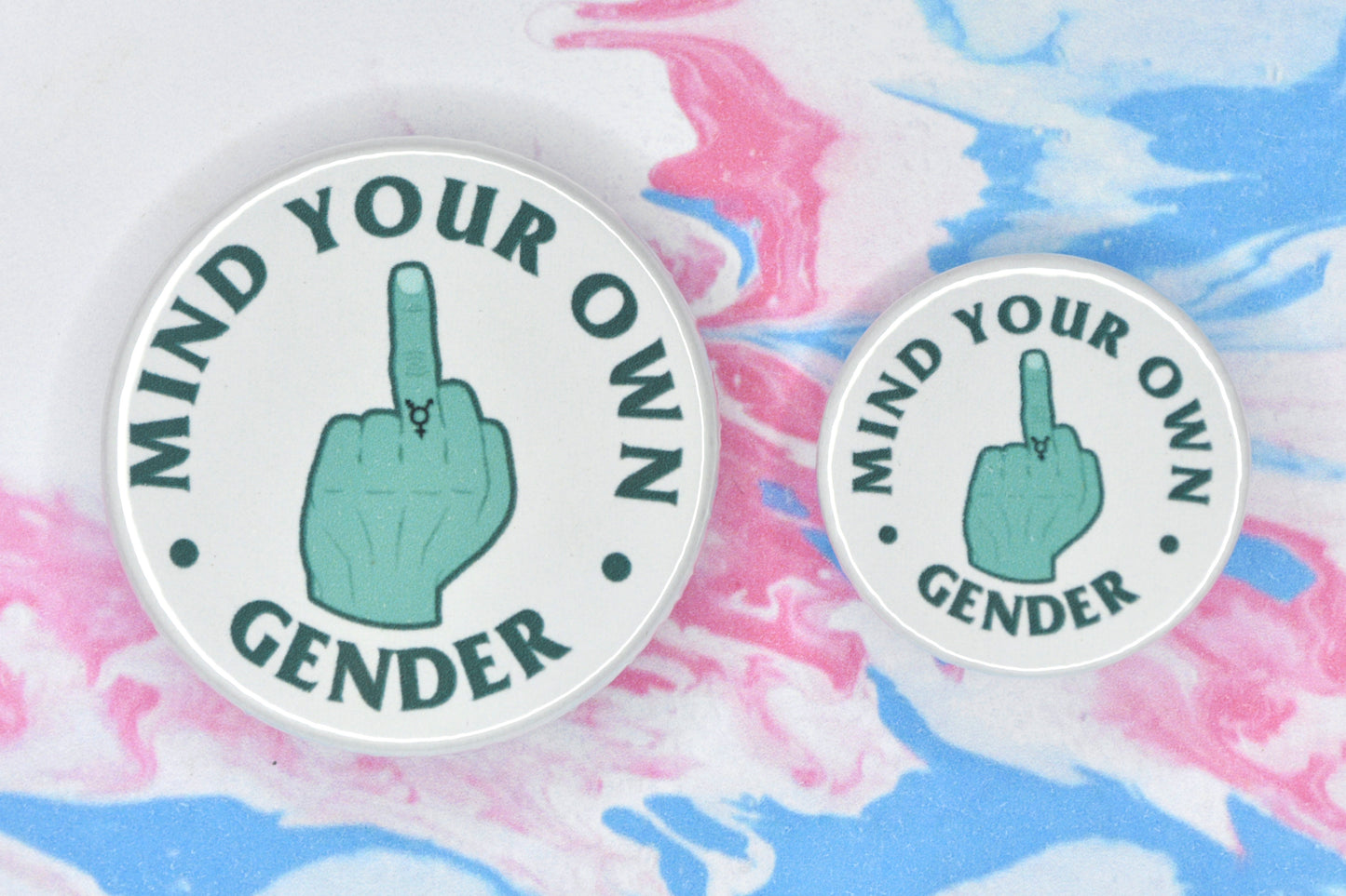 Two badges in sizes medium and small respectively, each featuring a white background with an illustration of a hand giving the middle finger in the centre. The hand is teal in colour and has a trans symbol tattoo on the middle finger. There is text around the illustration in a darker teal shade which reads "mind your own gender".