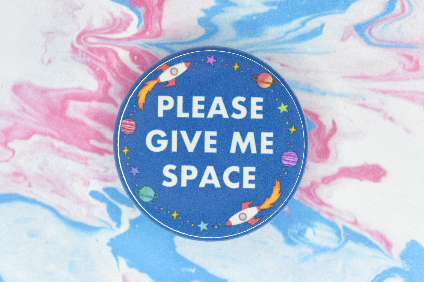 A hidden disability badge featuring a blue background and white text reading "Please give me space". There are space related illustrations surrounding the text including rockets, planets and stars.