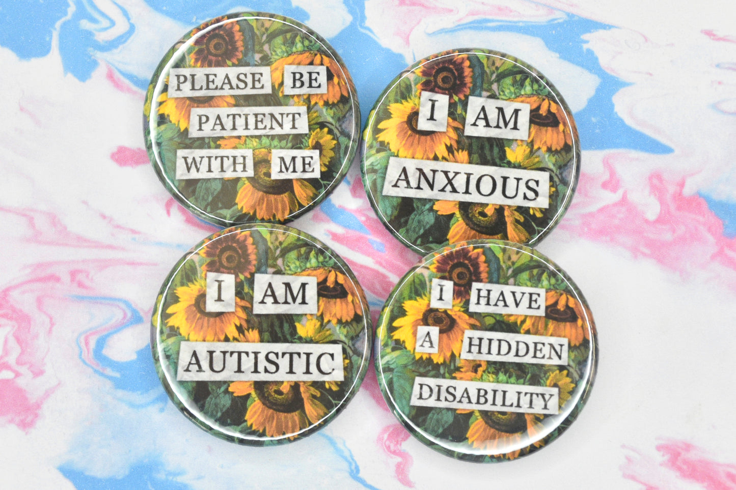 A collection of four circular button badges featuring a hidden disability sunflower design. Collaged text is overlaid on a painting of some sunflowers. The text reads "Please be patient with me", "I am anxious", "I am autistic" and "I have a hidden disability" respectively.