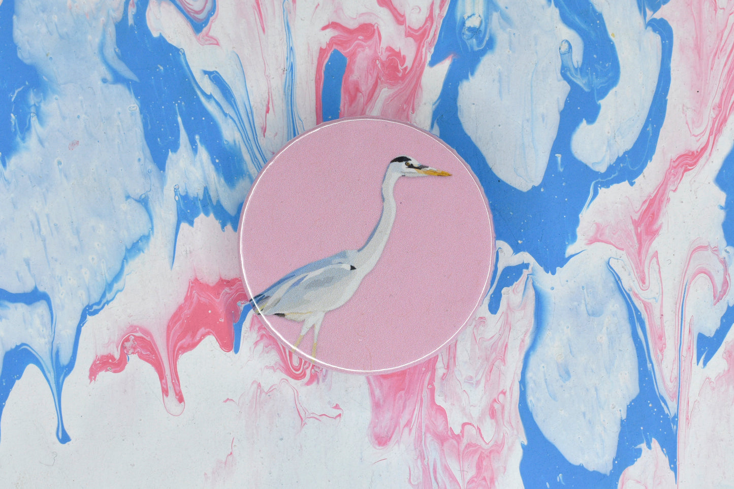 A circular pin badge with a pastel pink background featuring an illustration of a heron, a bird with grey and white feathers, a long neck and a yellow beak.