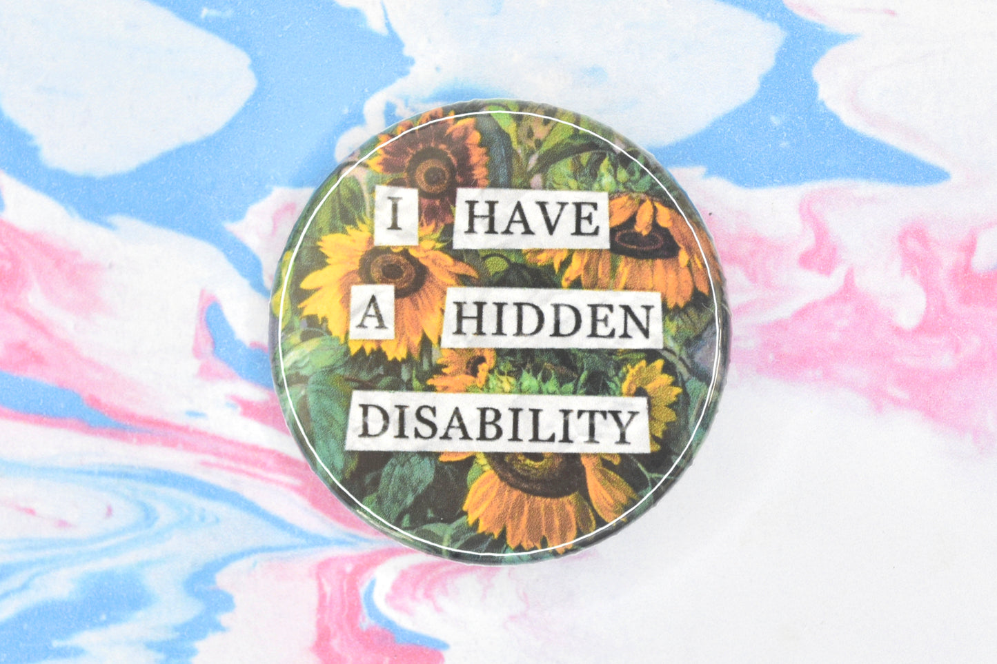 A circular button badge featuring a hidden disability sunflower design. Collaged text is overlaid on a painting of some sunflowers. The text reads "I have a hidden disability".