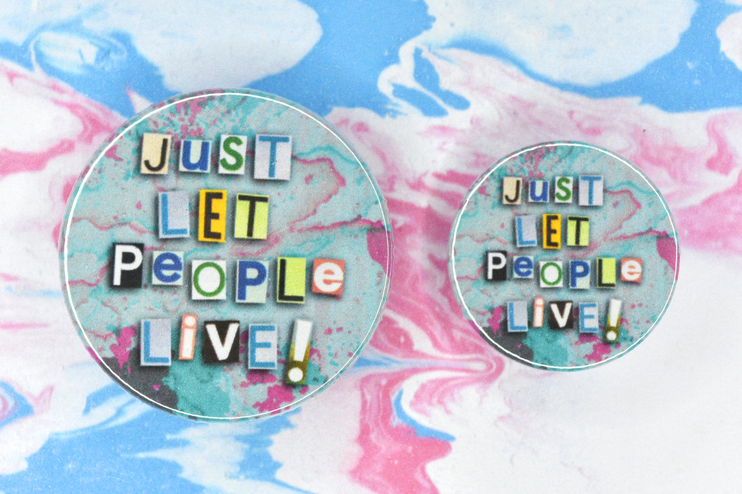 Button badges in two sizes featuring an LGBTQ+ pride collage design. Text reads "Just Let People Live!". The background colours of pink, white and blue are reminiscent of the trans pride flag.