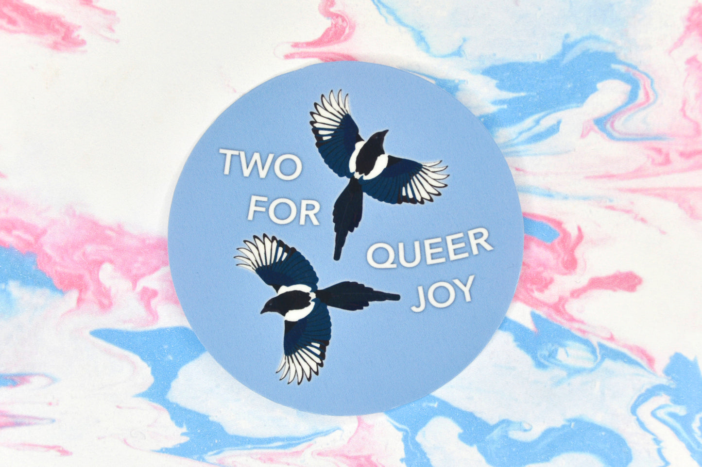 A circular LGBTQ+ pride vinyl sticker featuring an illustration of two magpies with text reading "two for queer joy"