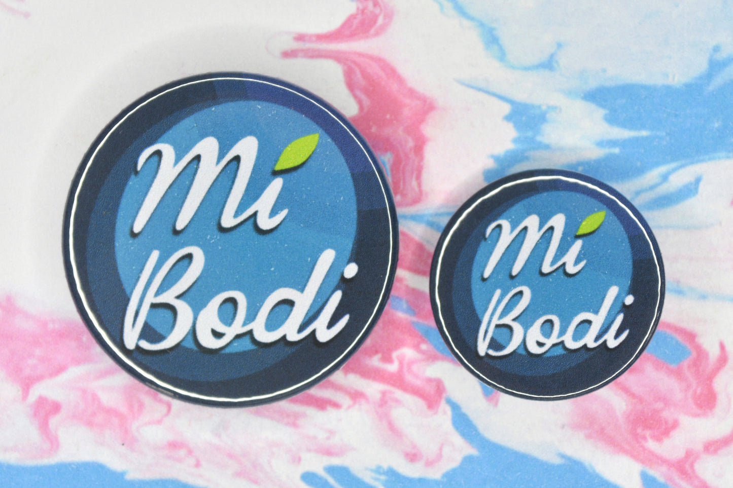 Two badges in sizes medium and small respectively, each featuring a background made up of different blue shades and white text reading "mi bodi" with a green leaf over the letter I in "Mi"