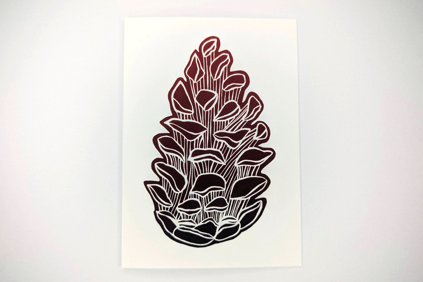 A greeting card featuring a lino print design of a pine cone printed in red ink on a white background.