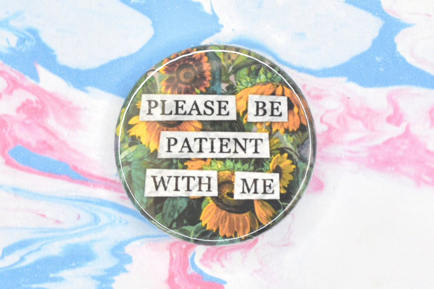 A circular button badge featuring a hidden disability sunflower design. Collaged text is overlaid on a painting of some sunflowers. The text reads "Please be patient with me".