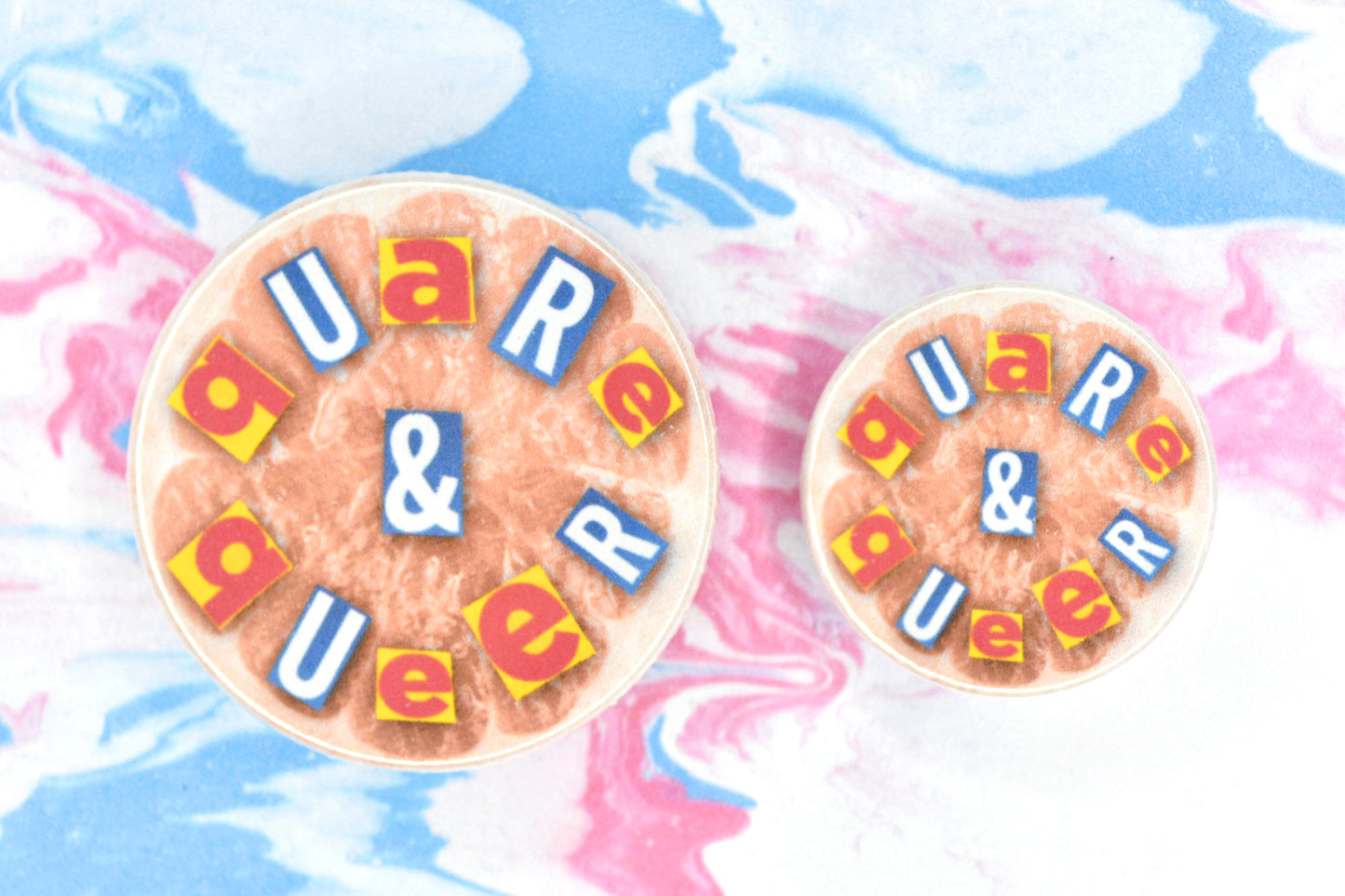 Button badges in two sizes featuring an LGBTQ+ pride design. Cutout text spells out "Quare & Queer". This is overlaid on a cutout image of a grapefruit with an orange filter applied.