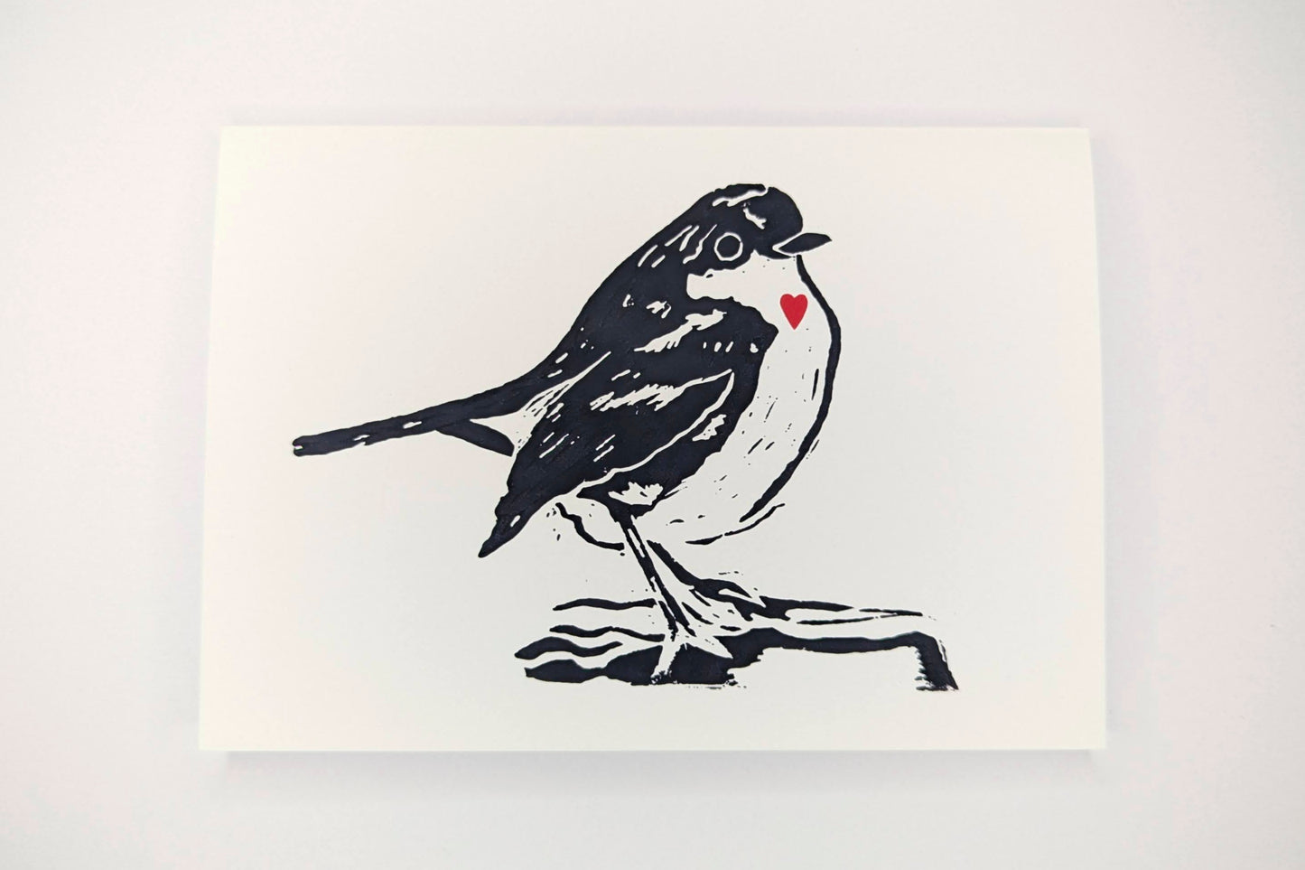 A tent fold greeting card featuring a lino print design of a robin printed in black ink on a white background with a small red heart over the bird's chest