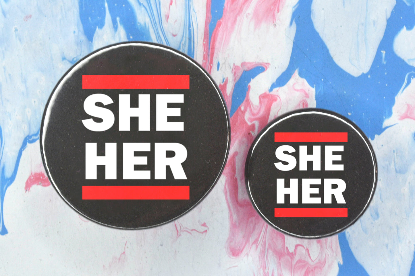 Two circular pin badges in sizes medium and small each with a black background, two rows of bold white upper case text and a solid red line above and below the text. The top row reads "she" and the bottom row reads "her".