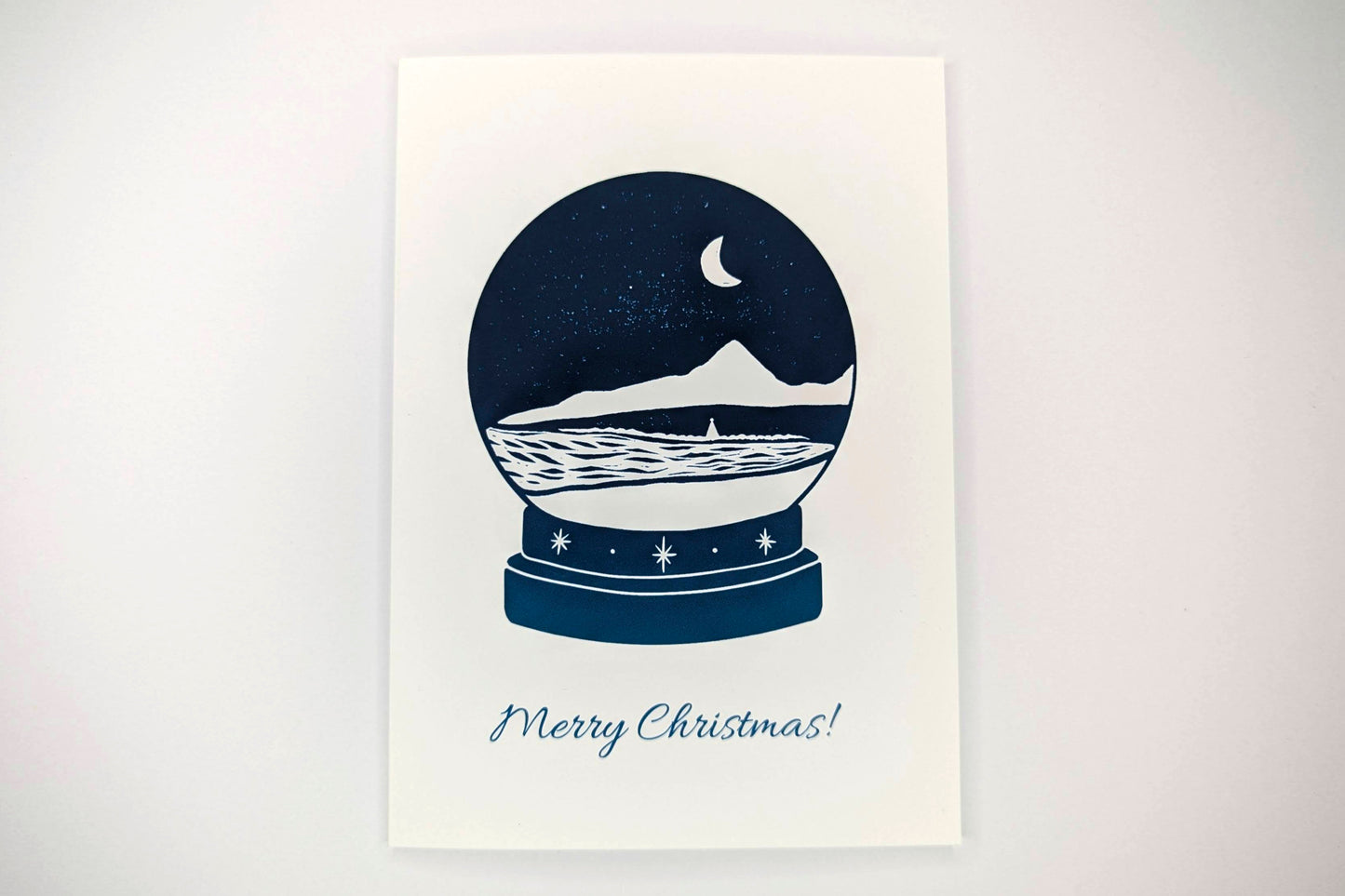 A greeting card featuring a lino print design of a snow globe with a coastal scene inside. Text below the snow globe reads "Merry Christmas"