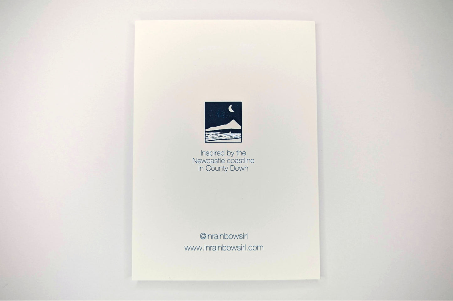 The back of the snow globe card which shows a close-up image of the design in the front and text reading "Inspired by the Newcastle coastline in County Down". Text at the bottom gives the shop's social media handle and website.
