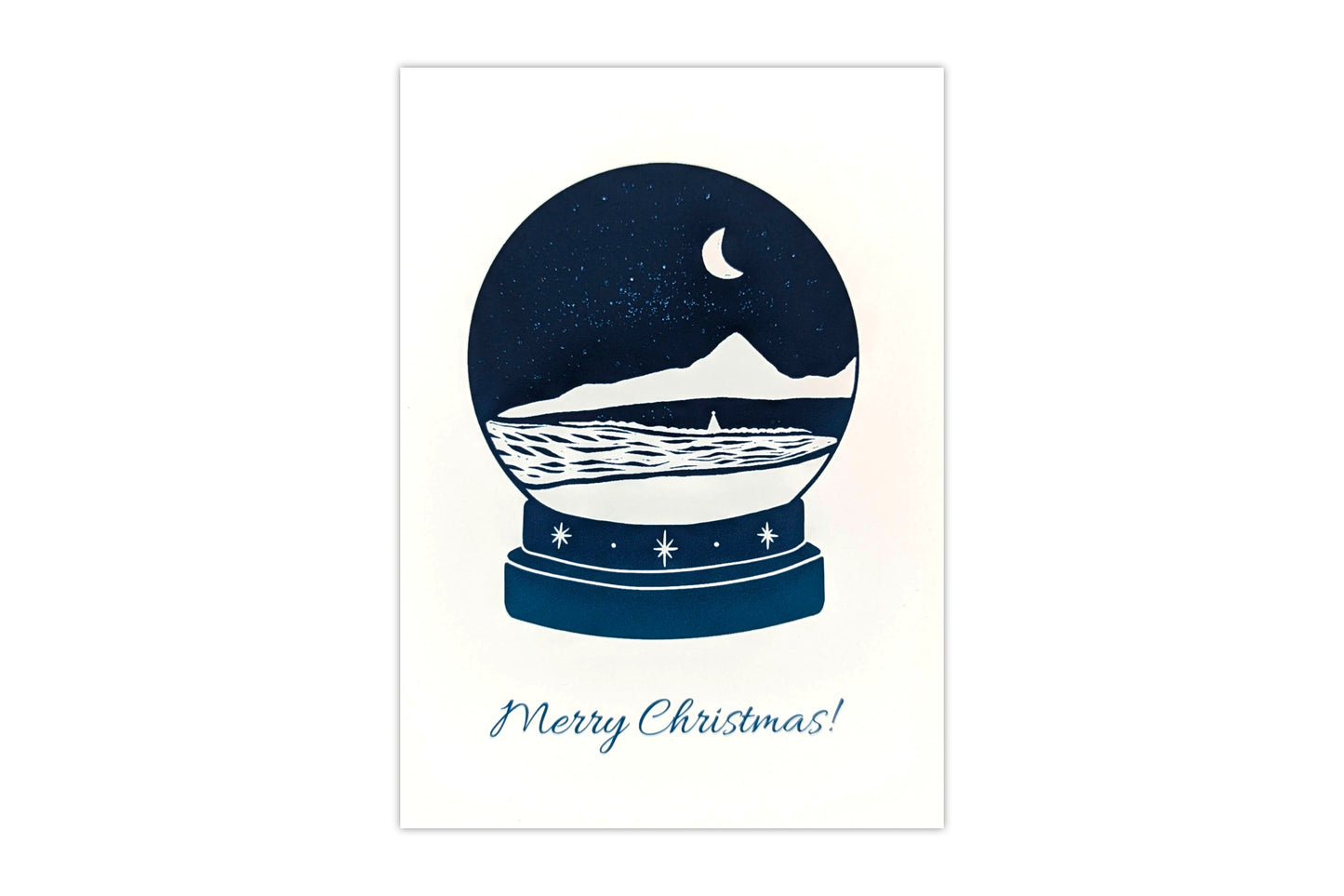 A greeting card featuring a lino print design of a snow globe with a coastal scene inside. Text below the snow globe reads "Merry Christmas"