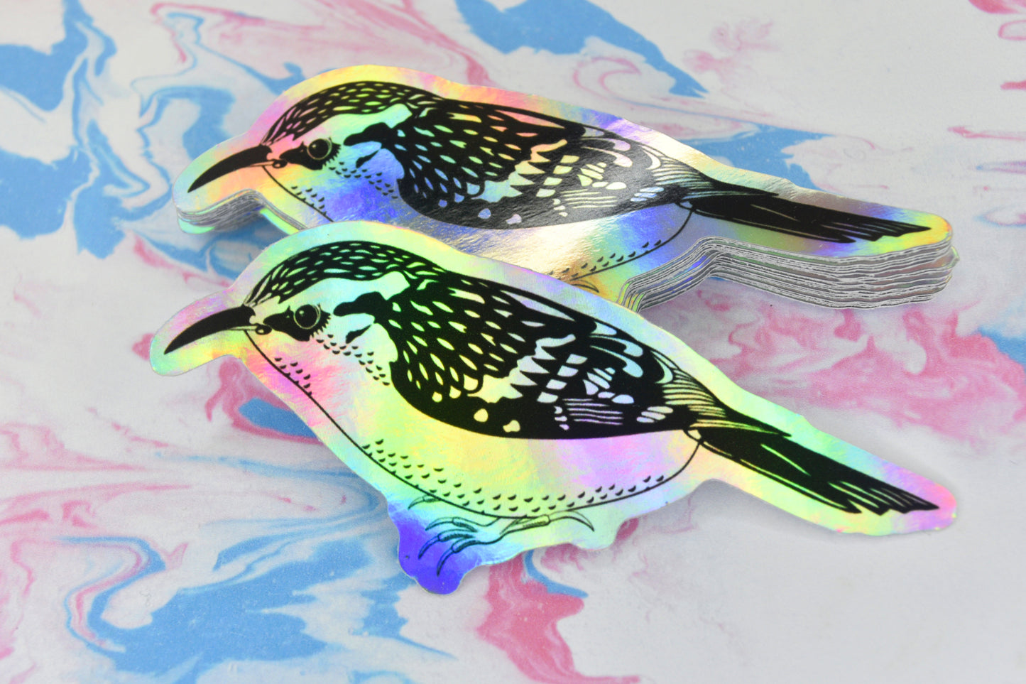 Some stickers featuring a treecreeper bird illustration in black ink on an iridescent holographic background. The holographic effect produces a metallic rainbow of colour across the surface of the sticker.