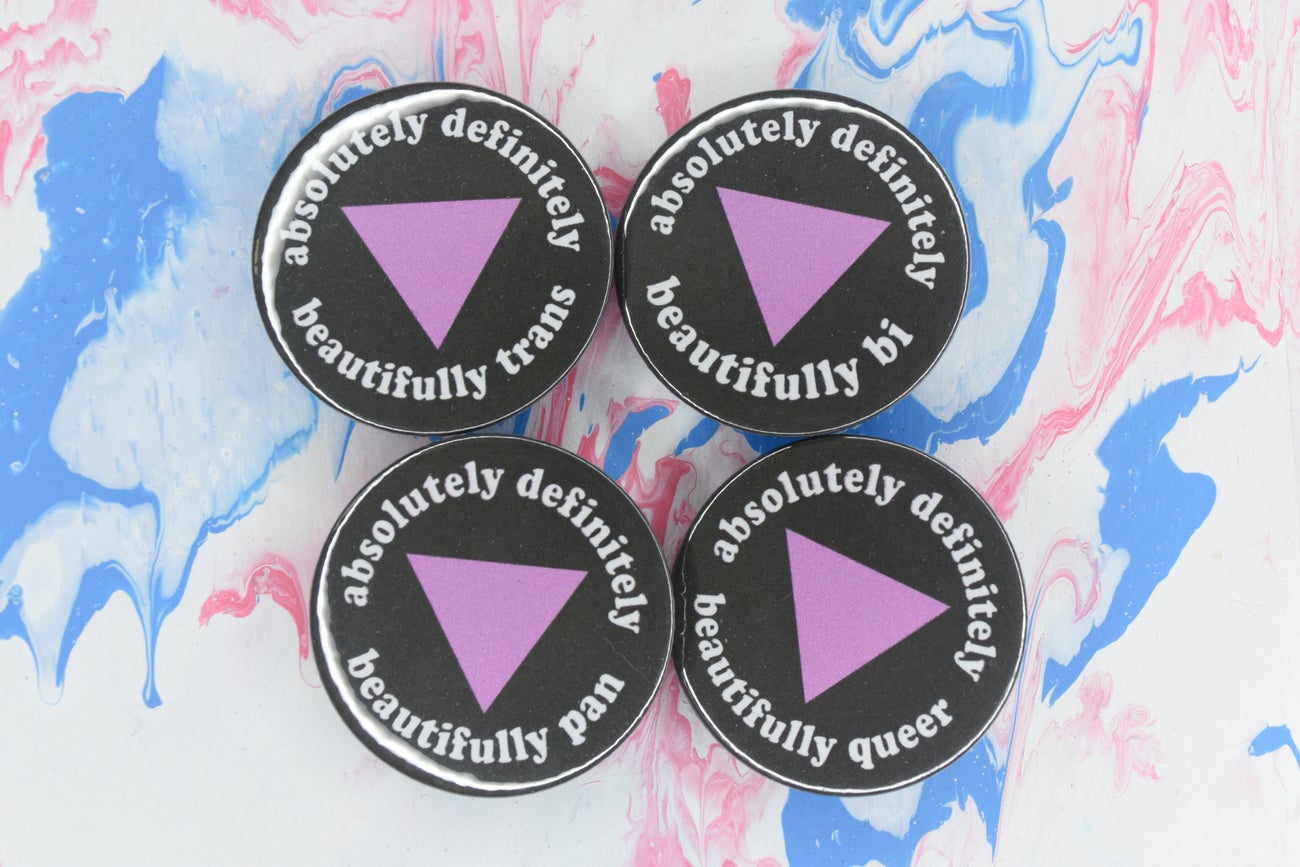 Four pin badges in size medium, all featuring a black background with a pink triangle in the centre and white text around the outside reading "absolutely definitely beautifully trans", "absolutely definitely beautifully bi", "absolutely definitely beautifully pan" and "absolutely definitely beautifully queer" respectively.