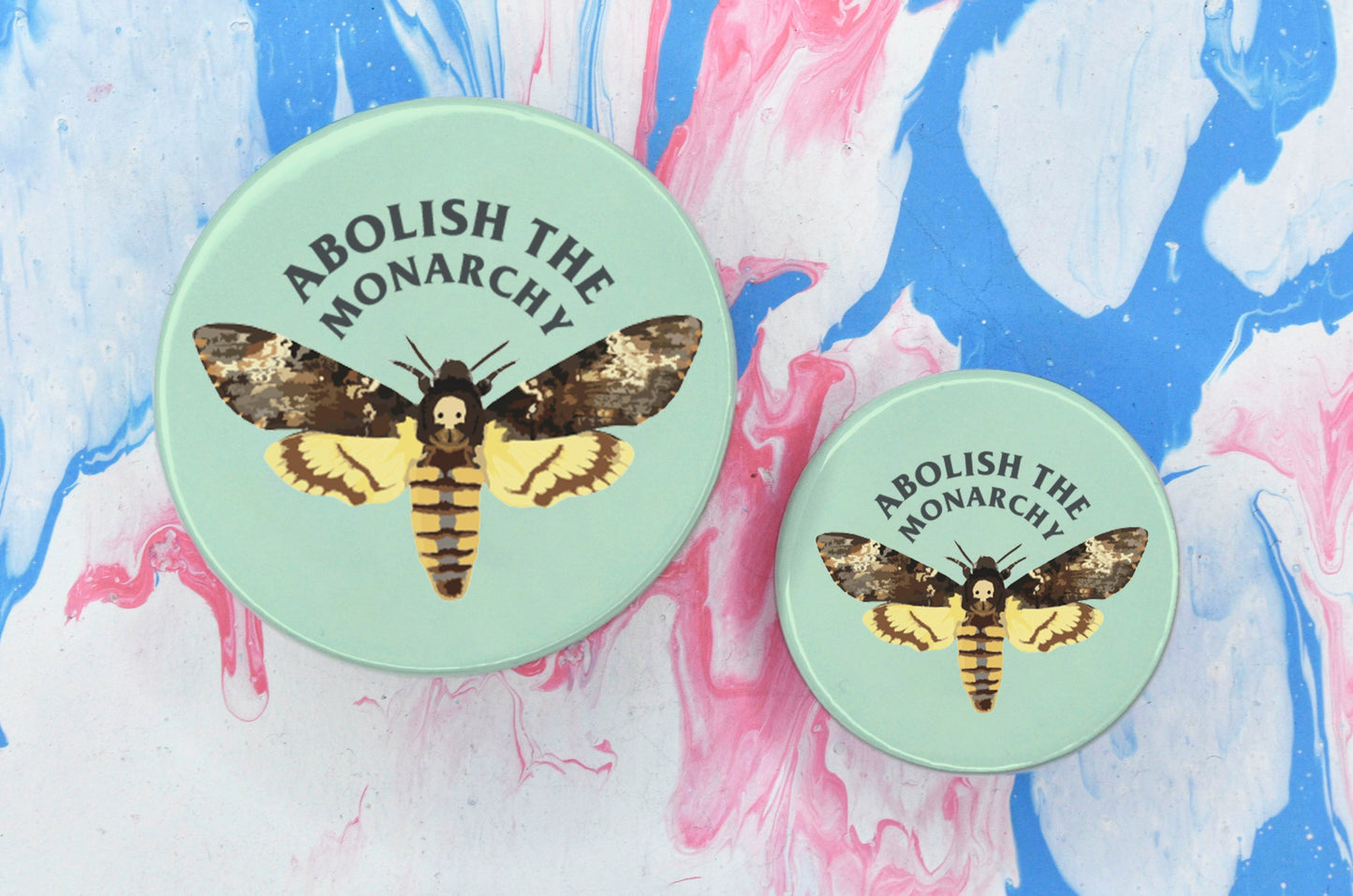 Two pin badges in sizes medium and small, both featuring an illustration of a moth on a light green background. There is text along the top of the badge reading "abolish the monarchy".