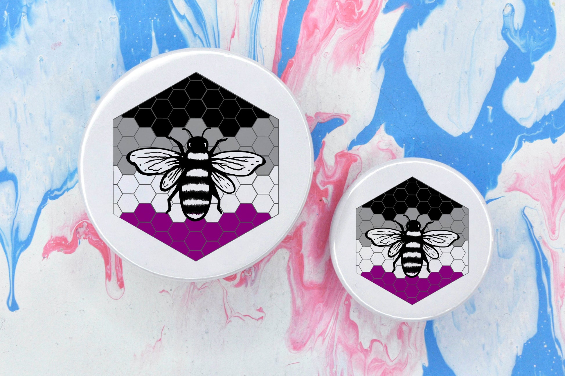 Two badges in sizes medium and small respectively, each featuring a white background with a black and white illustration of a bee overlaid on a bee hive pattern which is coloured in with the asexual pride flag colours (black, grey, white and dark pink).