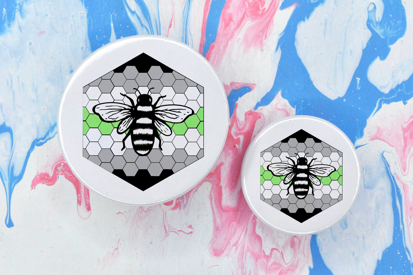 Two badges in sizes medium and small respectively, each featuring a white background with a black and white illustration of a bee overlaid on a bee hive pattern which is coloured in with the agender pride flag colours (black, grey, white and green).