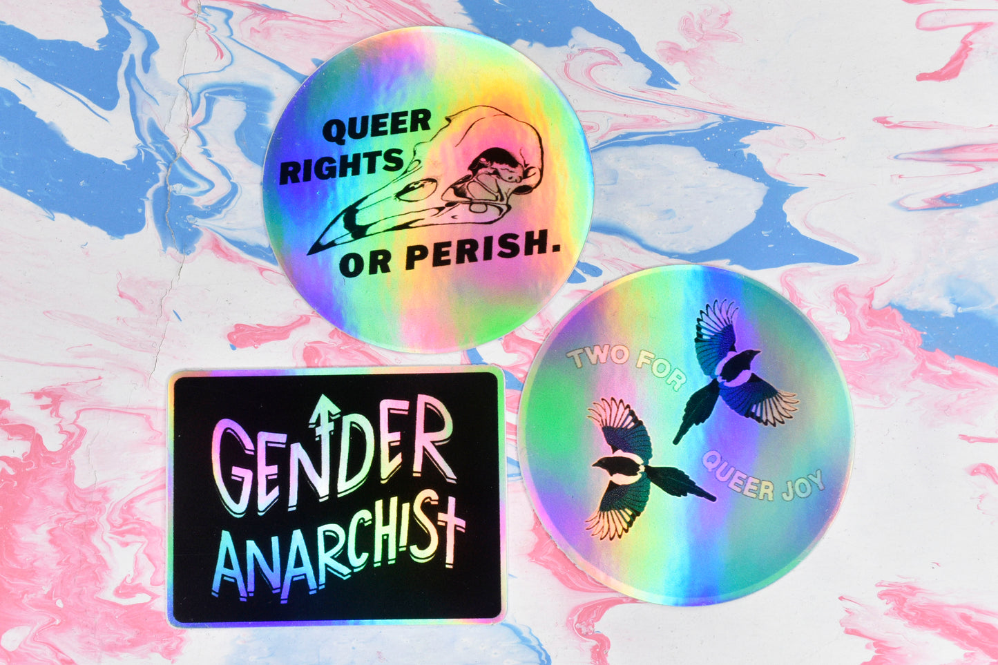 A photo of three holographic stickers. There is a round sticker featuring a bird skull illustration with text reading "queer rights or perish", a round sticker featuring an illustration of two magpies and text reading "two for queer joy" and a rectangular sticker with a black background and white/holographic text reading "Gender Anarchist".