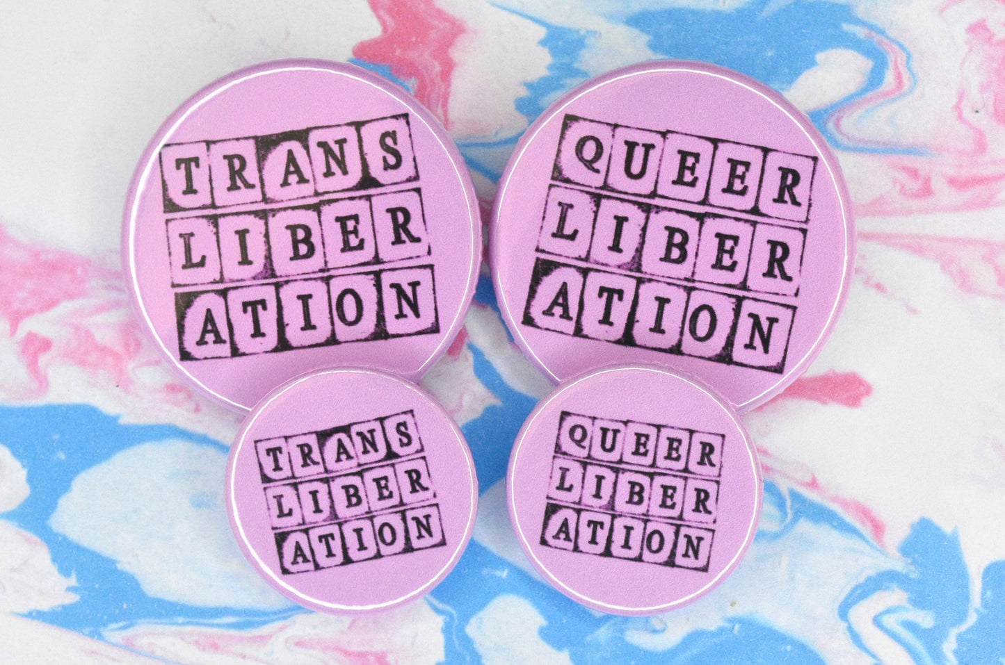 Four badges, two in size medium and two in size small, each featuring a pink background with text in a hand-stamped font reading "queer liberation" or "trans liberation".