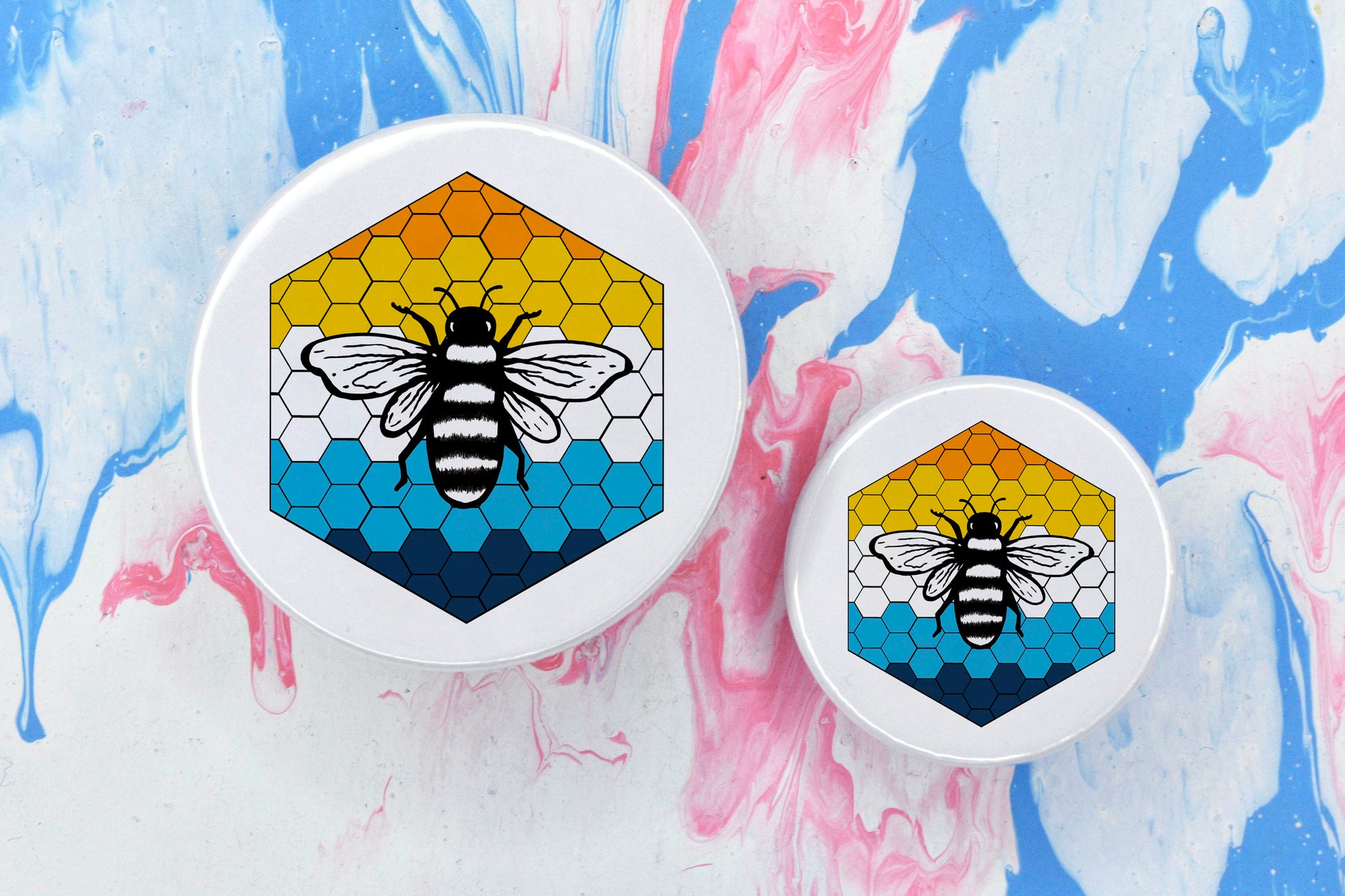 Two badges in sizes medium and small respectively, each featuring a white background with a black and white illustration of a bee overlaid on a bee hive pattern which is coloured in with the aroace pride flag colours (orange, yellow, white and blue).