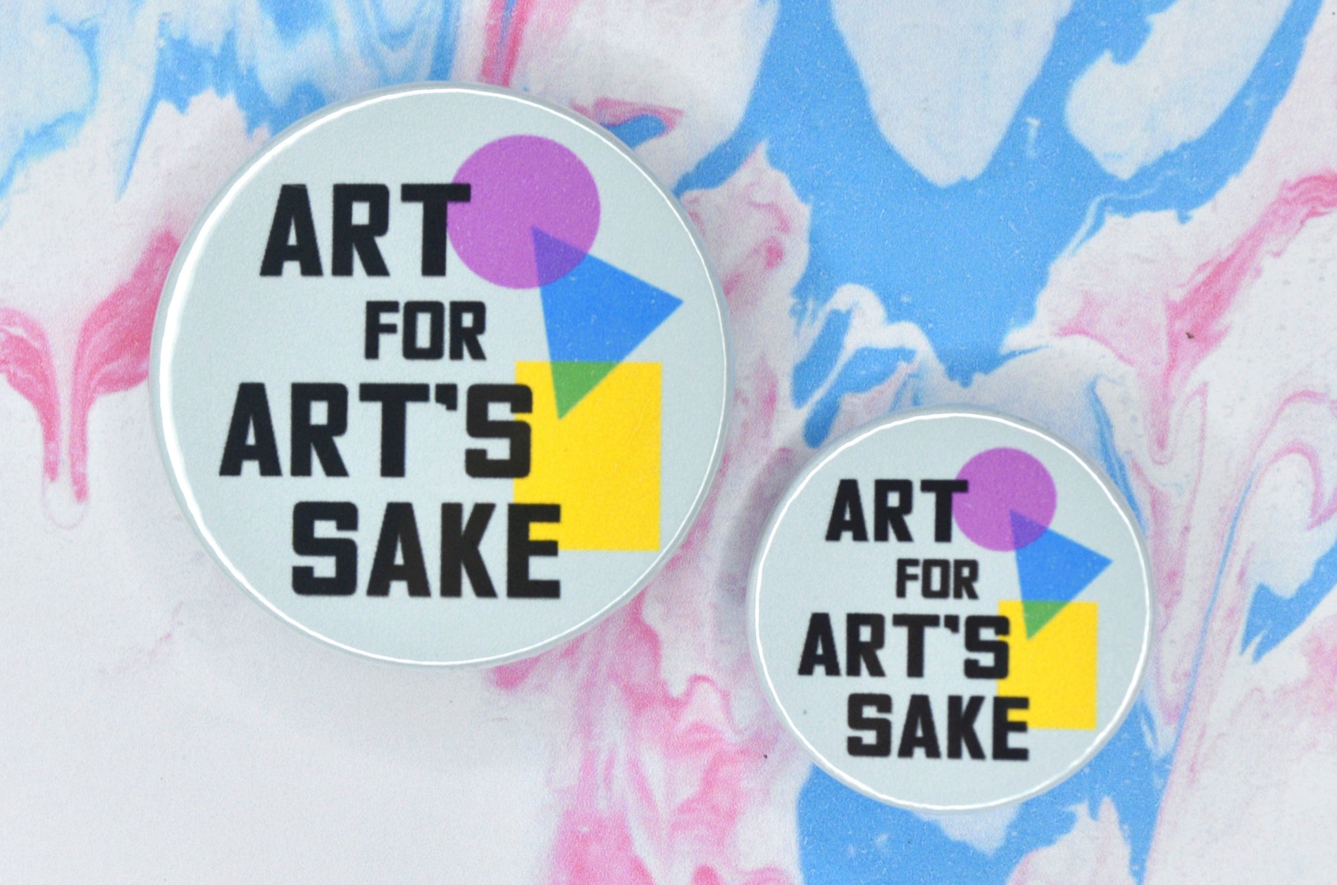 Two badges in sizes medium and small respectively, each featuring a pale blue background and black uppercase text reading "art for art's sake". There is a geometric design featuring a pink circle, blue triangle and yellow rectangle which are overlapping on the right hand side of the badges.