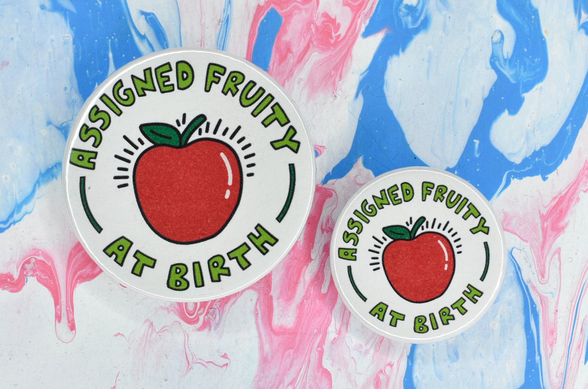 Two pin badges in sizes medium and small, both consisting of a white background with a red apple illustration in the centre. Light green text surrounds the apple reading "assigned fruity at birth".