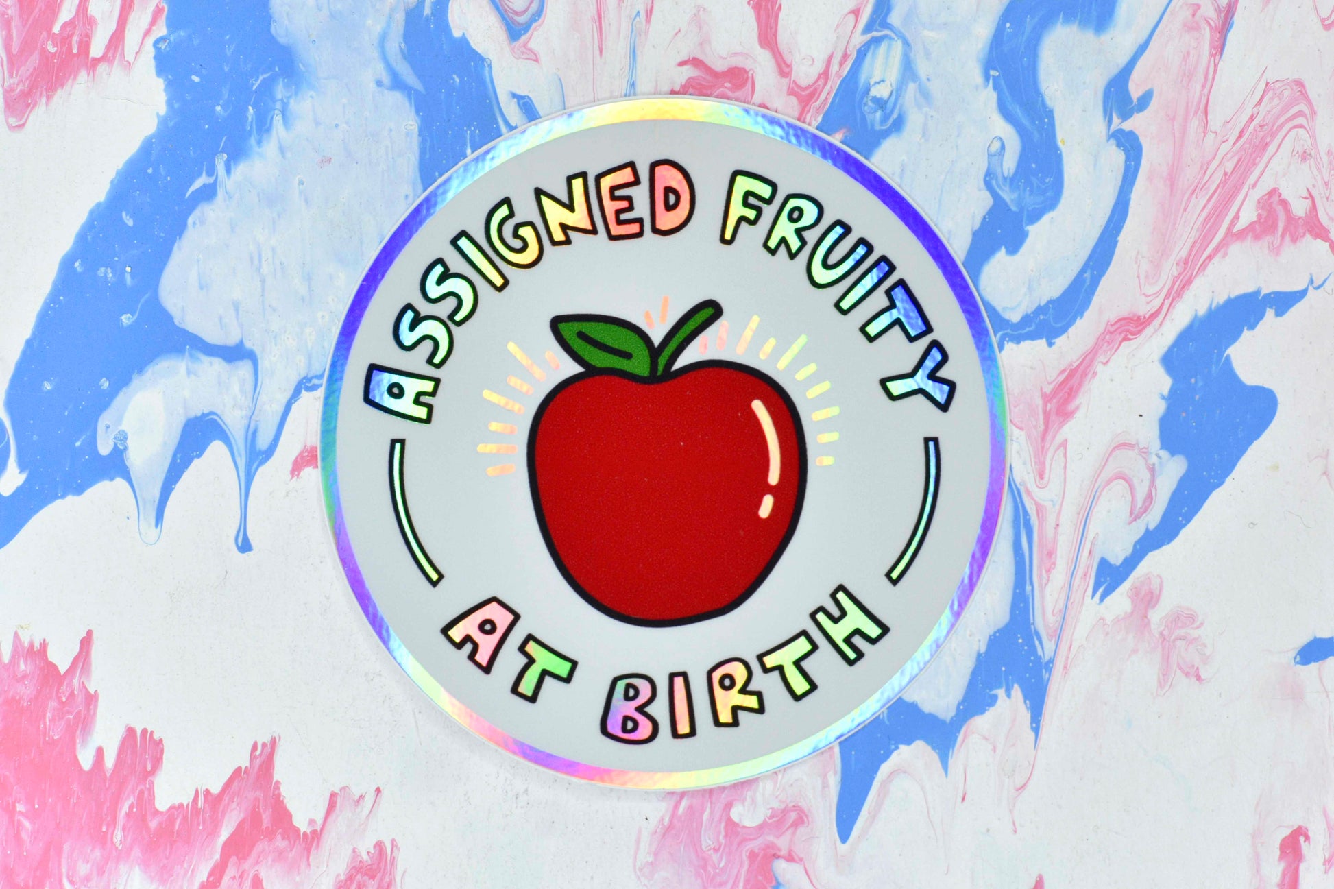 A circular sticker consisting of a white background with a red apple illustration in the centre. Holographic iridescent text surrounds the apple reading "assigned fruity at birth". The border of the sticker is also holographic.