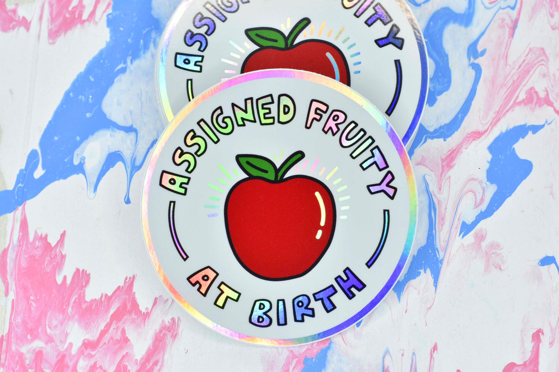 A circular sticker consisting of a white background with a red apple illustration in the centre. Holographic iridescent text surrounds the apple reading "assigned fruity at birth". The border of the sticker is also holographic.