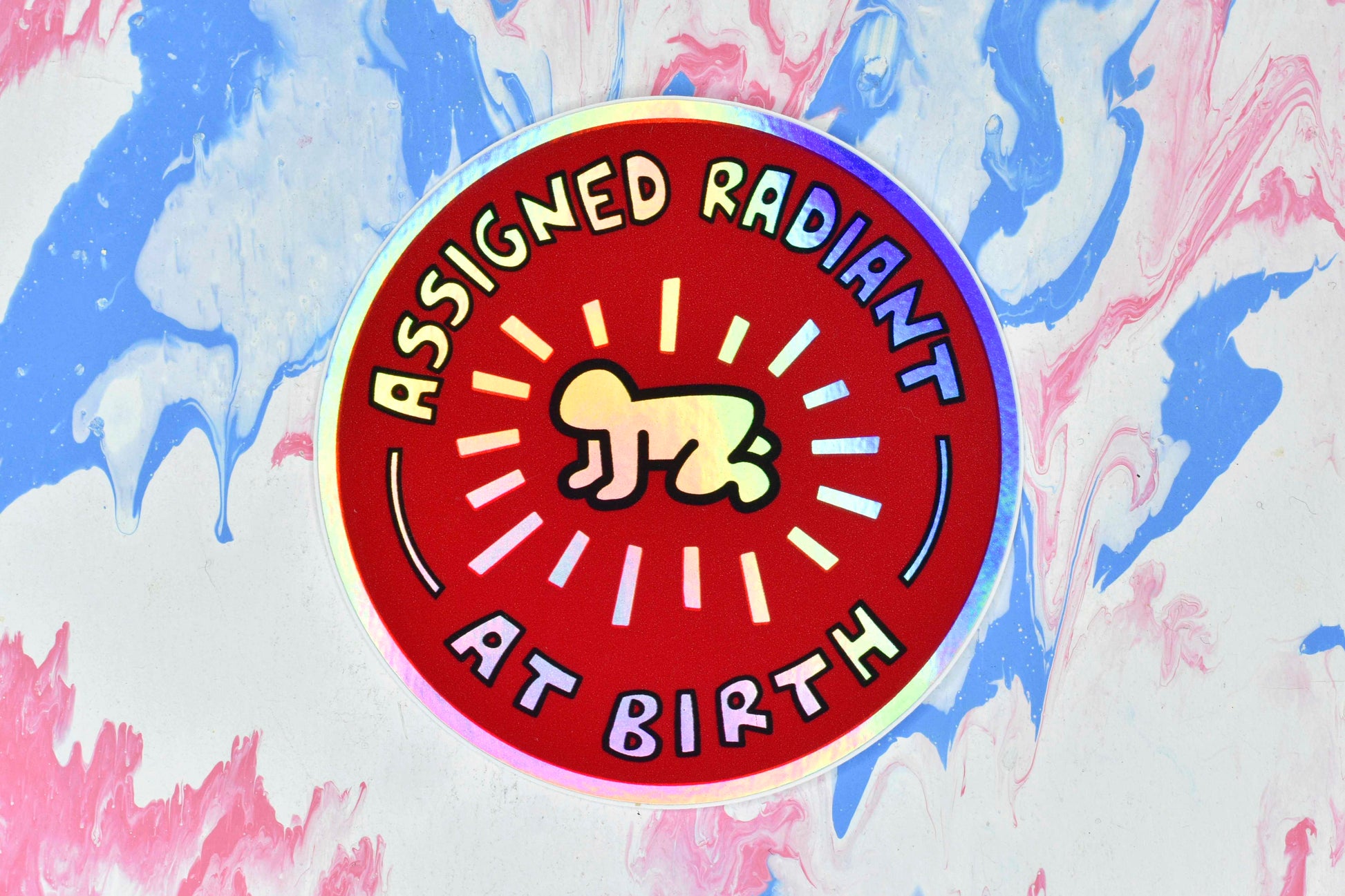 A circular sticker with a red background and Keith Haring's radiant baby illustration in the centre in black and white. There is text around the outside reading "assigned radiant at birth". The baby and text have a holographic finish that creates a rainbow effect across the metallic silver surface of the sticker paper.