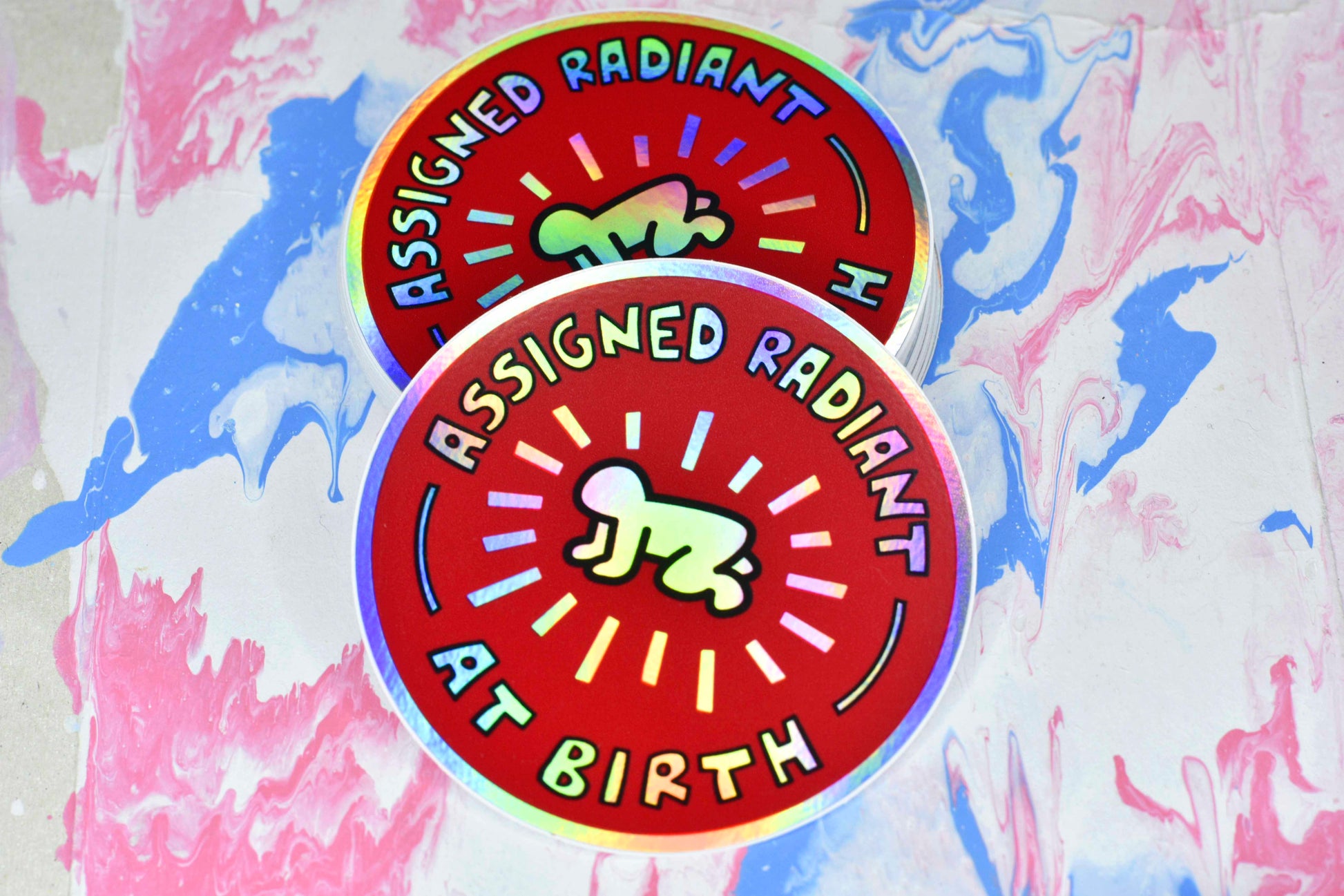 A circular sticker with a red background and Keith Haring's radiant baby illustration in the centre in black and white. There is text around the outside reading "assigned radiant at birth". The baby and text have a holographic finish that creates a rainbow effect across the metallic silver surface of the sticker paper.