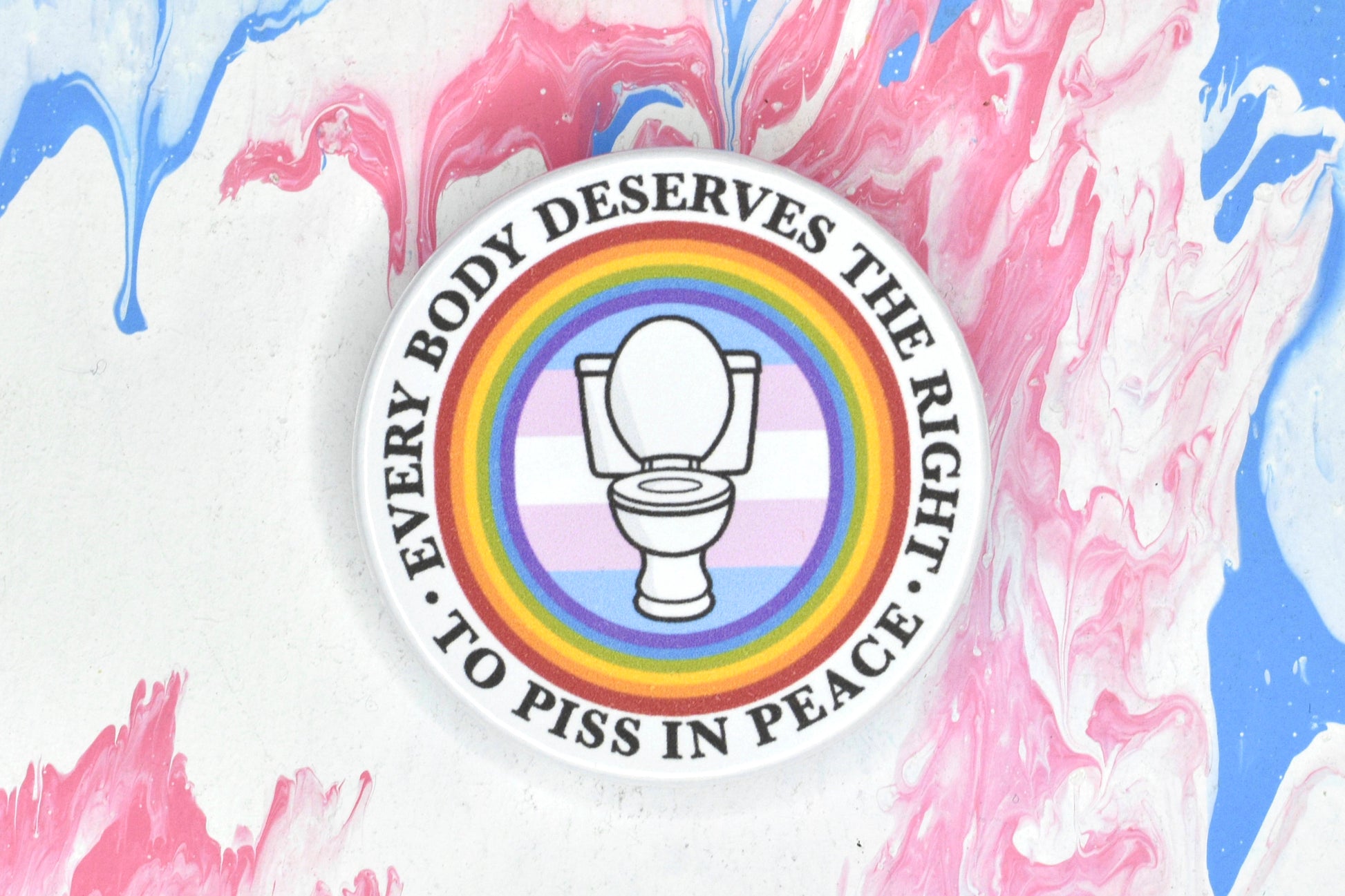 A circular button badge featuring a design depicting a white toilet in front of a trans pride flag with a rainbow circle around it. There is text running along the outside of the circle reading "every body deserves the right to piss in peace".