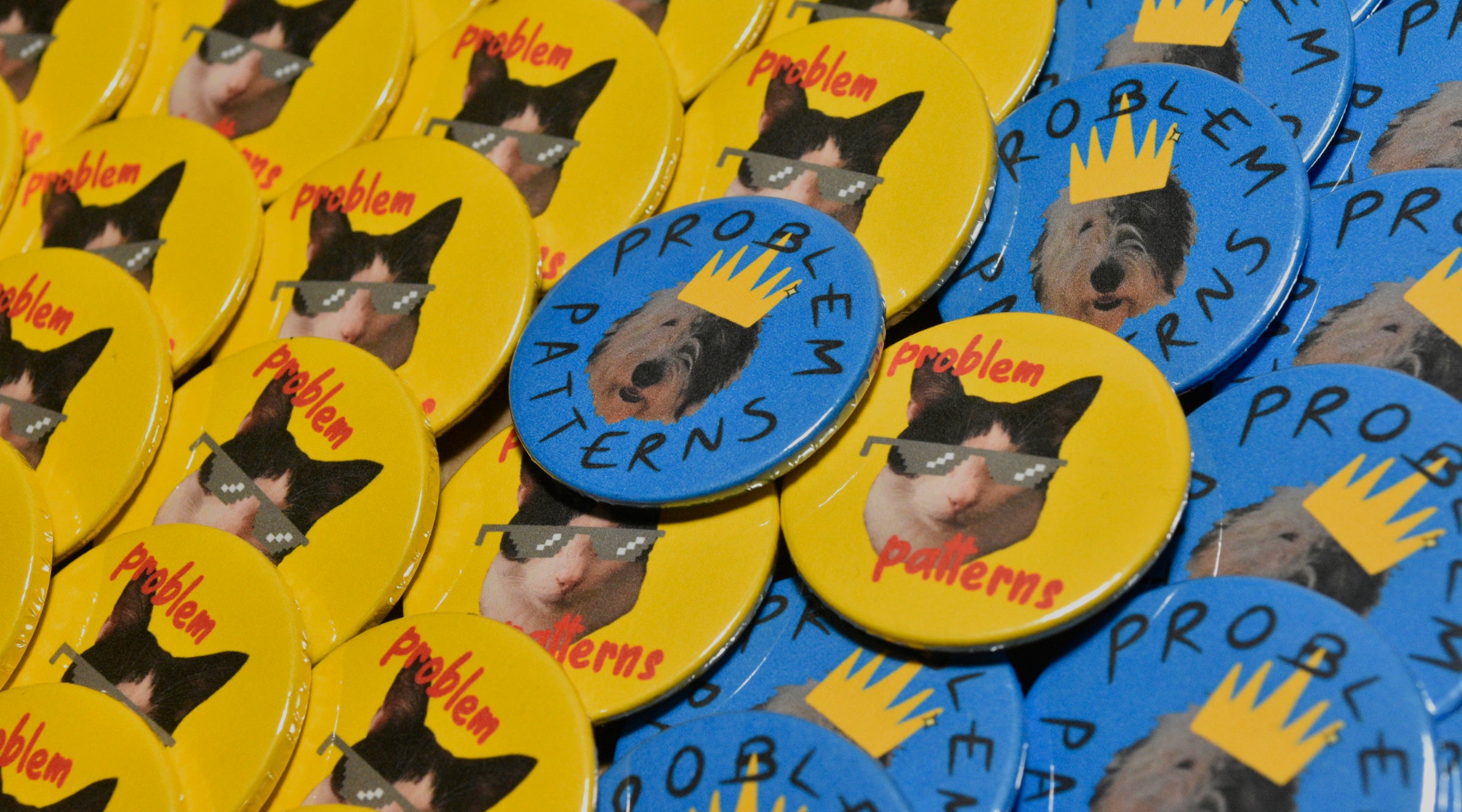 A large pile of badges for the band Problem Patterns in two designs. One design features a yellow background and a black and white cat wearing sunglasses with red text reading "Problem Patterns". The other features a blue background and a black and white old English sheepdog wearing a yellow crown with black text in a circle reading "Problem Patterns".