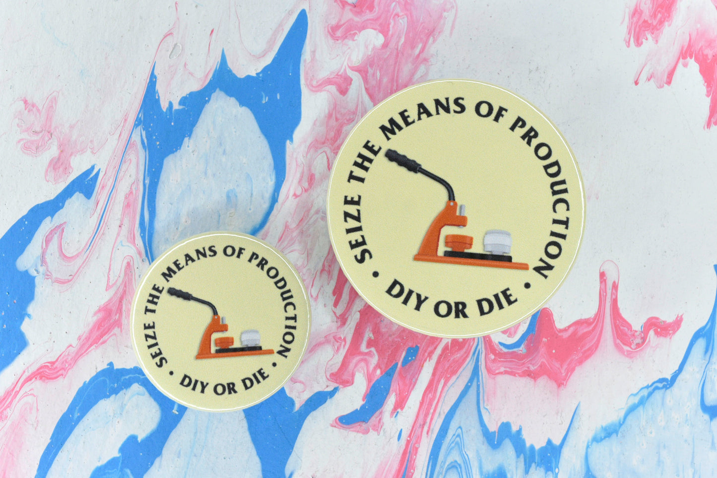 Two circular pin badges in sizes medium and small, each with a yellow background, an illustration of an orange badge making machine, and uppercase black serif text reading "seize the means of production - DIY or die".