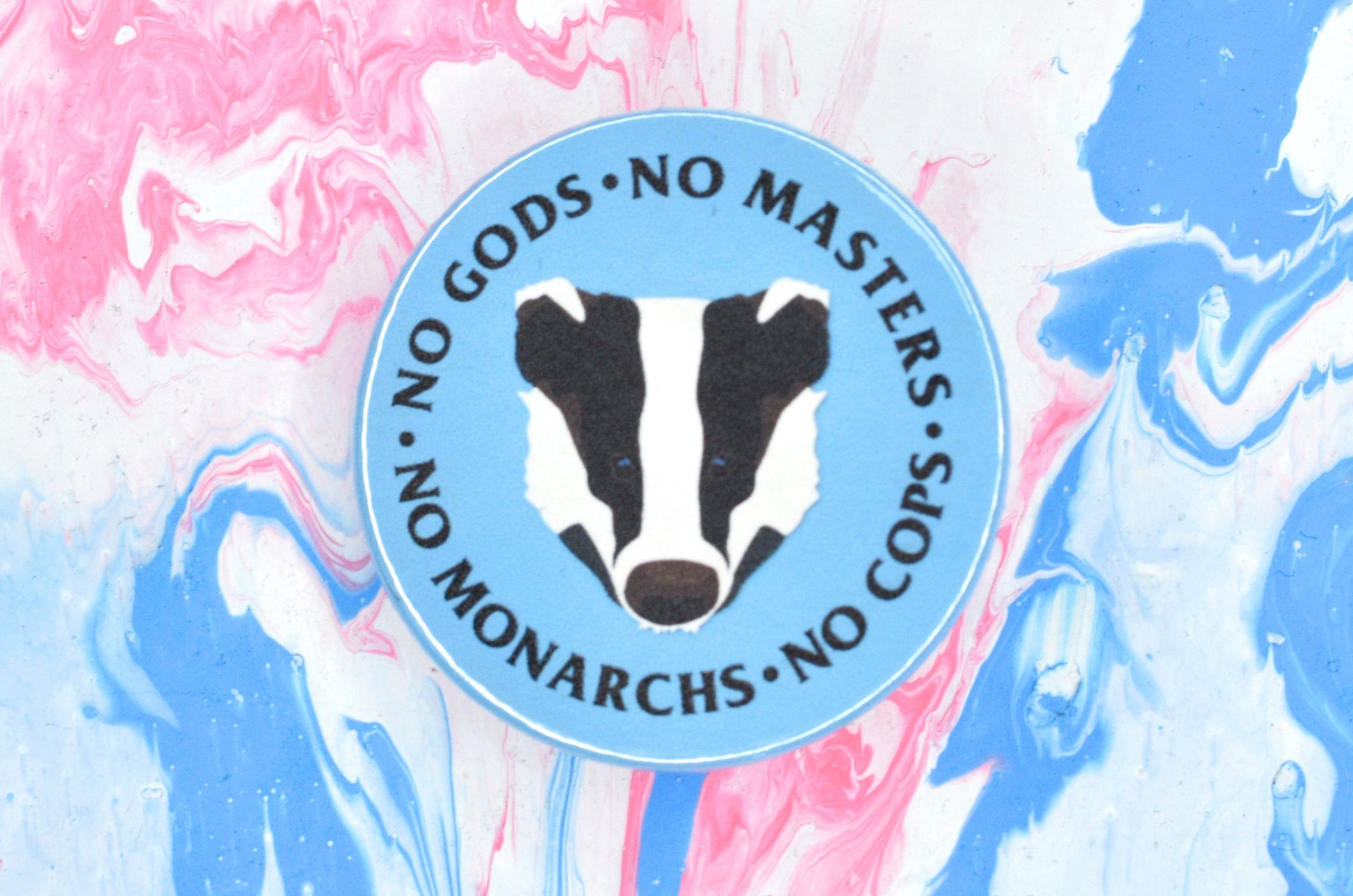 A circular pin badge with a light blue background and an illustration of a badger's face in the centre. There is black text around the outside of the badge reading "no gods, no masters, no monarchs, no cops".