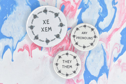 Three pin badges, one sized medium and the others sized small. Each badge has a white background and a circular border made up of small black and white illustrations of bees and pieces of foliage. There are two lines of black uppercase text inside the border of each badge. The medium badge reads "xe xem" while the smaller badges read "any pronouns" and "they them".