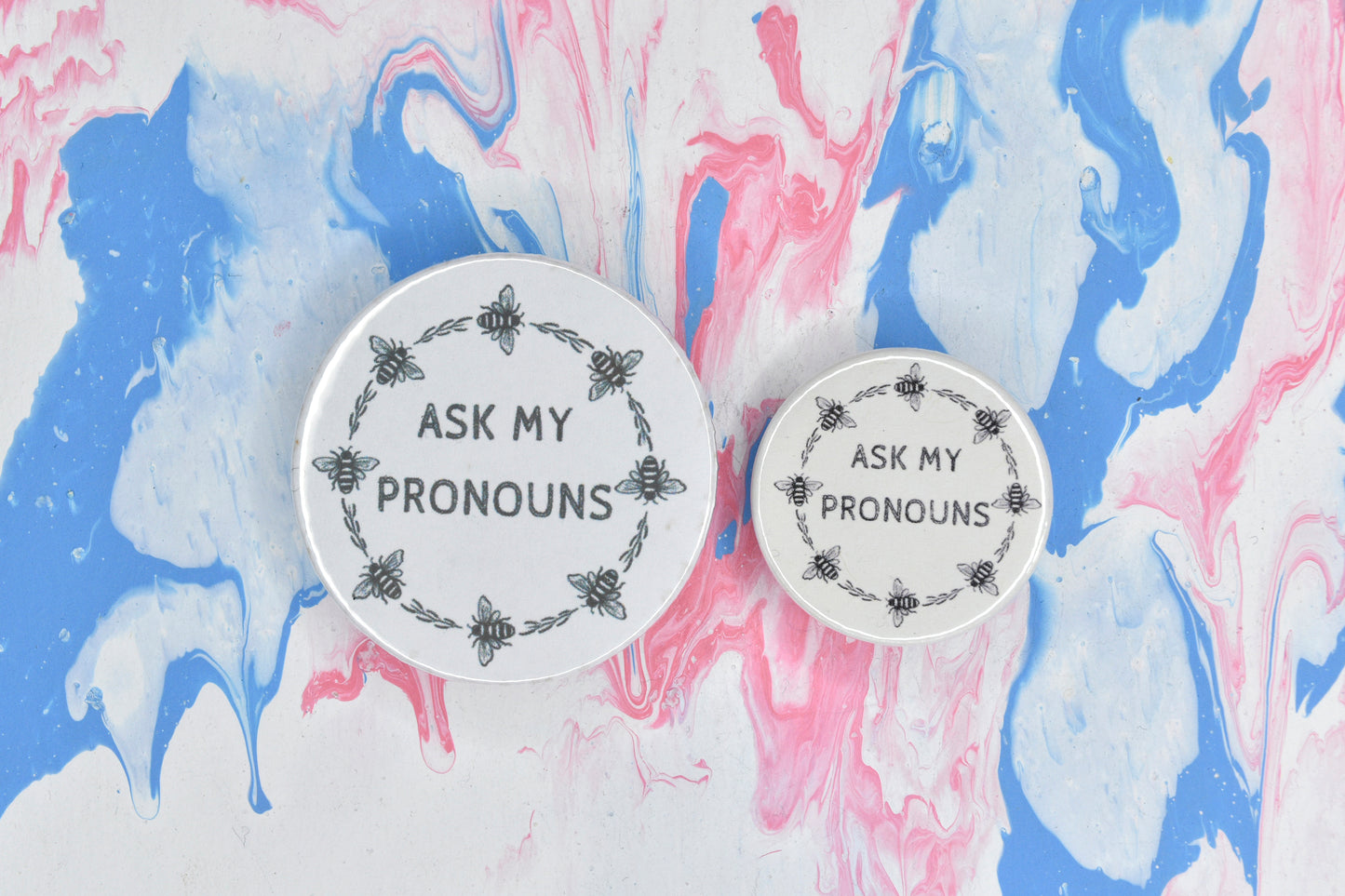 Two pin badges in sizes medium and small, both featuring a white background and a circular border made up of small black and white illustrations of bees and pieces of foliage. There are two lines of black uppercase text inside the border of each badge which reads "ask my pronouns".