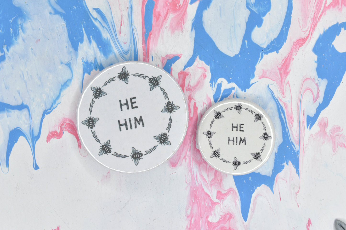 Two pin badges in sizes medium and small, both featuring a white background and a circular border made up of small black and white illustrations of bees and pieces of foliage. There are two lines of black uppercase text inside the border of each badge which reads "he him".