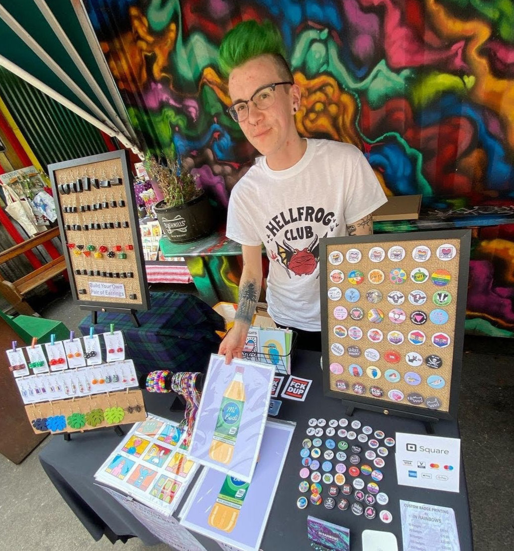 A photo of Alli, a white man in his 20s with green hair and glasses, standing behind his market table at Belfast Flea market with lots of art prints, badges and handmade jewellery on display