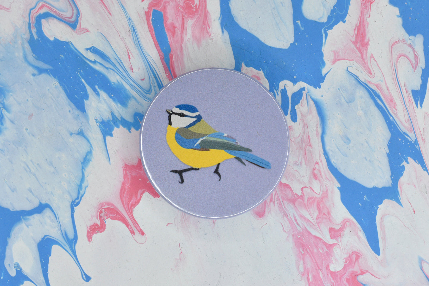 A circular pin badge with a pastel purple background featuring an illustration of a blue tit, a bird with blue, yellow, green and white markings.