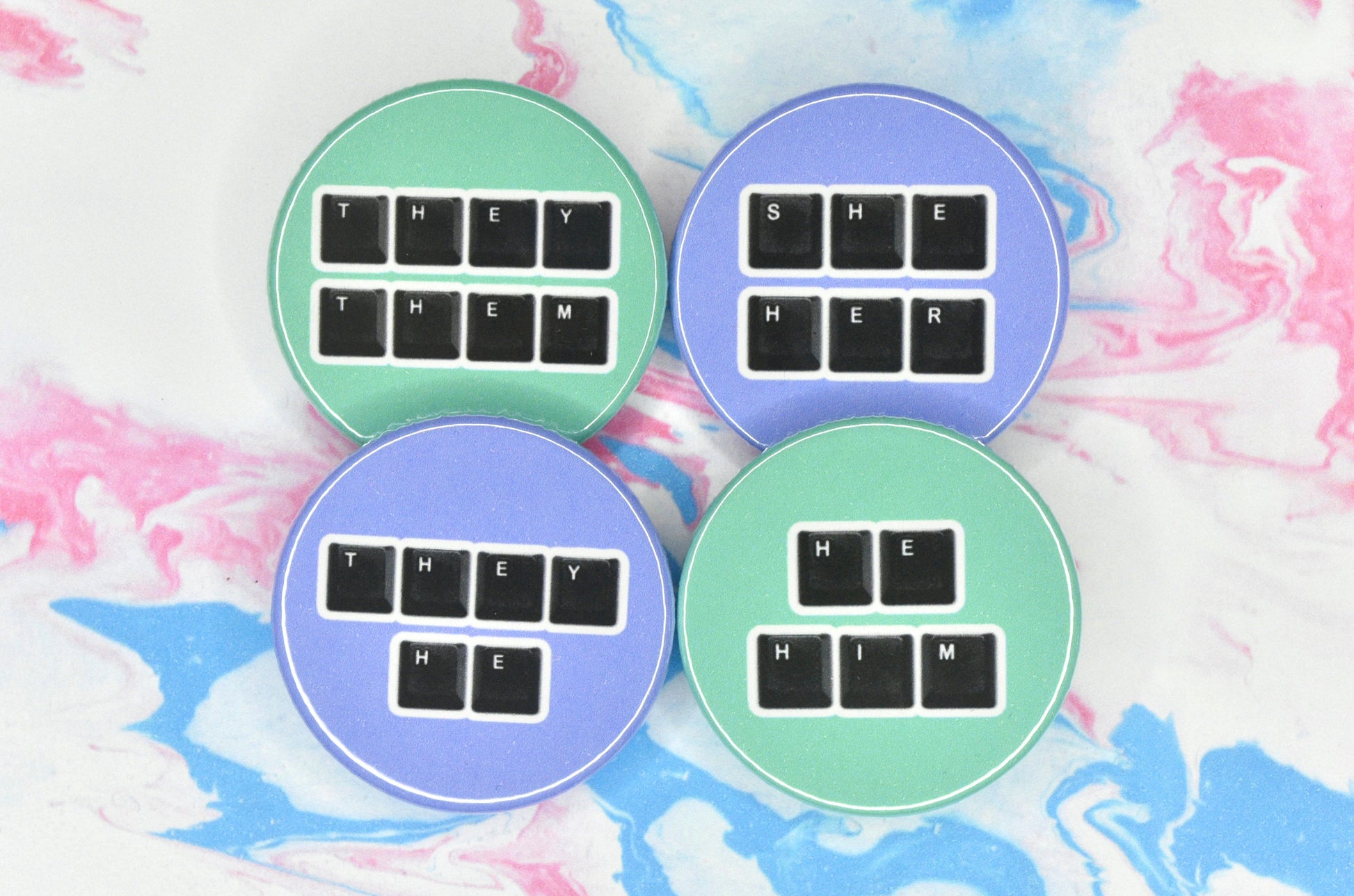 Four circular pin badges in size medium, two with a light purple background and two with a teal background, each featuring a set of pronouns spelt out using keyboard keys. The pronouns shown are "they/them", "she/her", "they/he" and "he/him".