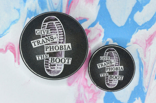 Two badges in sizes medium and small respectively, each featuring a dark grey background with a photograph of the bottom of a black combat boot. There is text overlaid on the photograph in a paper collage style which reads "give transphobia the boot".